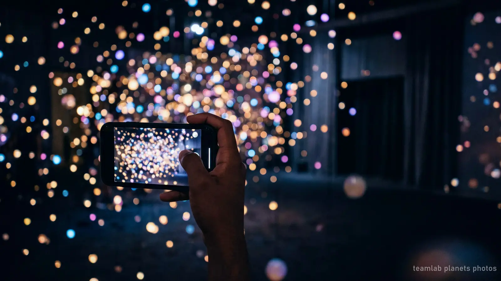 Illustration: a phone photographing a dark room of glowing coloured light, the kind of shot visitors take at teamLab Planets